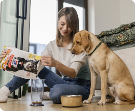Woman pouring Hill's Pet Nutrition food into a measuring container while a dog looks attentively.