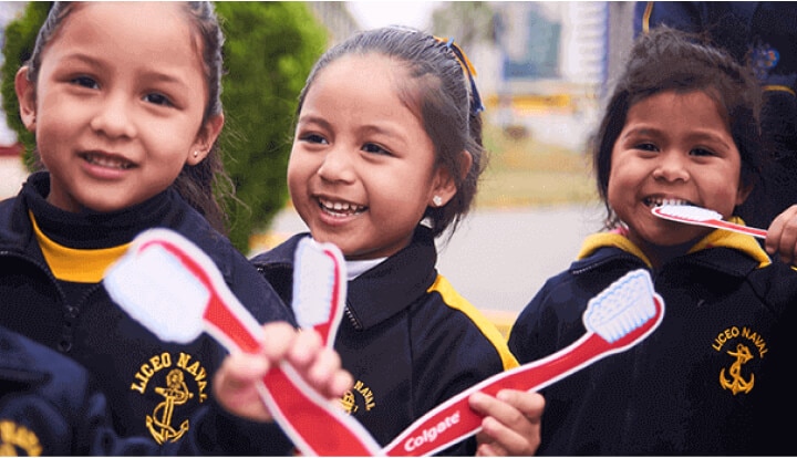 Three smiling young girls wearing school uniforms holding Colgate toothbrushes outdoors.