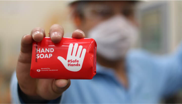 Person wearing a mask holds a red bar of hand soap labeled "HAND SOAP #Safe Hands" for cleaning and hygiene.