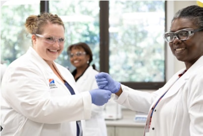 Two scientists in lab coats and gloves smile and shake hands in a laboratory setting.